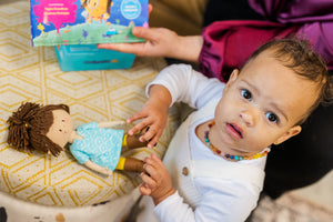 Little girl looking up into the camera while playing with Mini Ava Cultural Doll from Little Patakha with her blue long shirt, yellow pants and brown boots