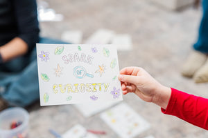 A kid's arm in a red sleeve holding a Little Patakha postcard in her hand that reads Spark Of Curiosity that she has colored in based on the number and color key provided