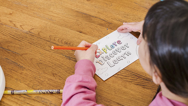 Looking at a girl in a pink sweatshirt as she colors a postcard with the words explore, discover and learn on it with other Little Patakha products like affirmation cards, books and stickers around her