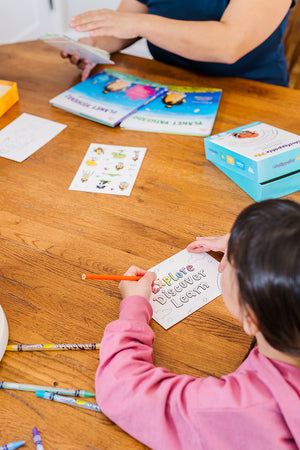 Looking at a girl in a pink sweatshirt as she colors a postcard with the words explore, discover and learn on it with other Little Patakha products like affirmation cards, books and stickers around her