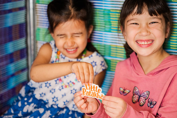 Two girls sitting in front of a shelf of books holding up a magnet of the Little Patakha logo. One girl in blue and white dress has her eyes closed and the other in a pink sweatshirt has her eyes open with both smiling